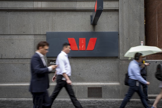People walk past a large, grey stone wall featuring a prominent red and white logo of a financial institution. The scene captures individuals in business attire, some holding umbrellas or wearing headphones.