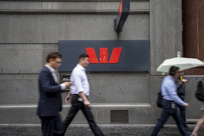 People walk past a large, grey stone wall featuring a prominent red and white logo of a financial institution. The scene captures individuals in business attire, some holding umbrellas or wearing headphones.