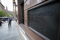 A bustling urban street scene features people walking past a building with a sign reading Commonwealth Bank of Australia. The area is lined with modern and historic architecture, with a clock tower visible in the distance. Trees add a touch of greenery, and a bus is seen on the street.