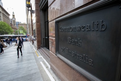 A bustling urban street scene features people walking past a building with a sign reading Commonwealth Bank of Australia. The area is lined with modern and historic architecture, with a clock tower visible in the distance. Trees add a touch of greenery, and a bus is seen on the street.