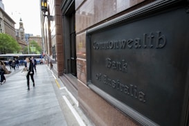 A bustling urban street scene features people walking past a building with a sign reading Commonwealth Bank of Australia. The area is lined with modern and historic architecture, with a clock tower visible in the distance. Trees add a touch of greenery, and a bus is seen on the street.