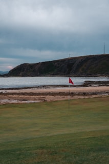 A green golf course with a red flag in the foreground, bordered by a sandy beach and rocky shoreline. Behind the water, there is a hilly landscape under an overcast sky.