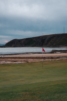 A green golf course with a red flag in the foreground, bordered by a sandy beach and rocky shoreline. Behind the water, there is a hilly landscape under an overcast sky.
