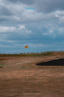 A golf course featuring a yellow flag planted on a pole in the middle of a green area. The grass varies in color, with some patches appearing dry and brown. There are two sand bunkers visible, one in the foreground and one in the distance. The background shows a cloudy sky with shades of blue and grey.