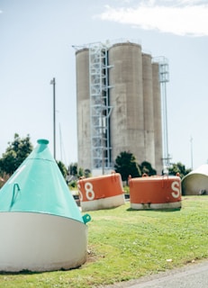 Concrete silos stand prominently in the background, framed by orange and turquoise maritime buoys scattered on the grass in front. The scene is set outdoors on a clear day, with some trees visible in the distance.