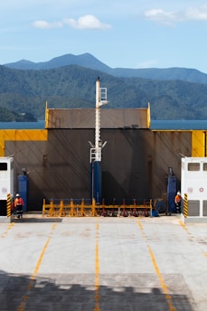 A ferry deck with two people in orange safety gear is seen, with a backdrop of green hills and a blue sky. The deck is open and spacious, with yellow and black safety markings and barriers. The ramp is raised, and there is a structure in the center that looks like a mast or pole.