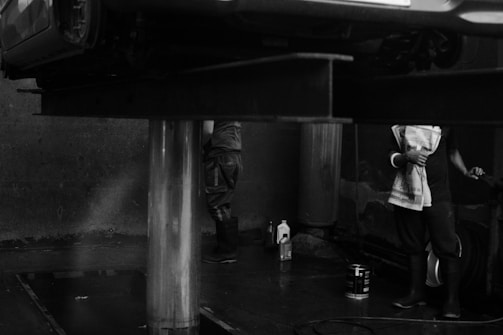 A monochromatic scene with a vehicle elevated on a hydraulic lift in what appears to be a garage or car service area. Two people are partially visible, one standing with arms crossed holding a towel, and another person wearing boots. Various containers, possibly lubricants or cleaning products, are on the floor.
