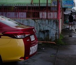 A red and yellow Toyota Vios car is parked on a wet street next to a green building with red metal fencing. A sign for drug testing is visible, and a person walks away in the distance wearing a black top and shorts.