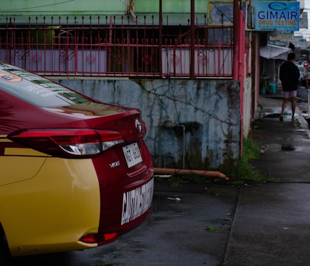 A red and yellow Toyota Vios car is parked on a wet street next to a green building with red metal fencing. A sign for drug testing is visible, and a person walks away in the distance wearing a black top and shorts.