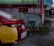 A red and yellow Toyota Vios car is parked on a wet street next to a green building with red metal fencing. A sign for drug testing is visible, and a person walks away in the distance wearing a black top and shorts.