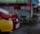 A red and yellow Toyota Vios car is parked on a wet street next to a green building with red metal fencing. A sign for drug testing is visible, and a person walks away in the distance wearing a black top and shorts.