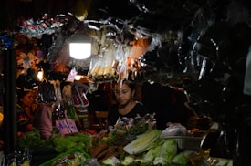 A dimly lit market stall with a variety of vegetables in the foreground, such as lettuce, carrots, and green peppers. Several hanging bags containing different items are visible, along with a bright overhead lamp illuminating the scene. People are present behind the stall, partially obscured by the merchandise.