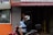 A person wearing a helmet and black hoodie is riding a red motorbike with a blue towel over their lap. An older person stands in a doorway to the right, partially obscured by shadows. The background features a storefront with signs for a physician and dental clinic. An orange and gray tiled facade is visible, along with a window revealing another person inside.