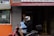A person wearing a helmet and black hoodie is riding a red motorbike with a blue towel over their lap. An older person stands in a doorway to the right, partially obscured by shadows. The background features a storefront with signs for a physician and dental clinic. An orange and gray tiled facade is visible, along with a window revealing another person inside.