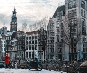 A European cityscape featuring a row of historic buildings with traditional architecture, including a tall clock tower. In the foreground, there is a canal with several parked bicycles and a person in a red coat standing nearby. The sky is overcast with scattered clouds, creating a slightly moody atmosphere.