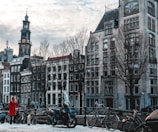 A European cityscape featuring a row of historic buildings with traditional architecture, including a tall clock tower. In the foreground, there is a canal with several parked bicycles and a person in a red coat standing nearby. The sky is overcast with scattered clouds, creating a slightly moody atmosphere.