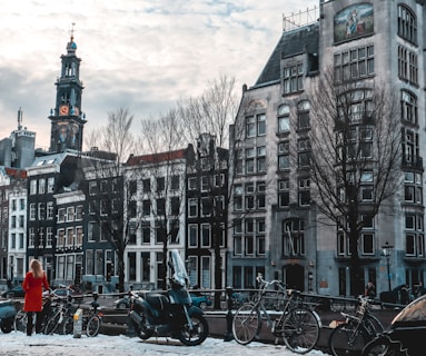 A European cityscape featuring a row of historic buildings with traditional architecture, including a tall clock tower. In the foreground, there is a canal with several parked bicycles and a person in a red coat standing nearby. The sky is overcast with scattered clouds, creating a slightly moody atmosphere.