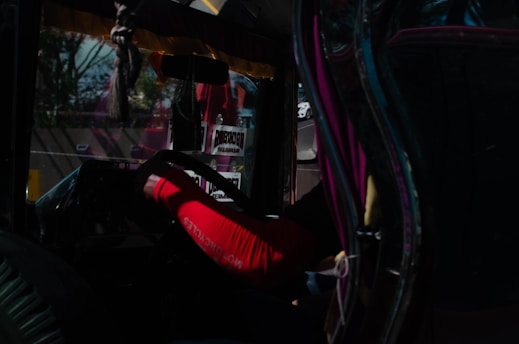 Inside a vehicle, a person wearing a red sleeve is holding the steering wheel. The interior is dimly lit with reflections and decals visible on the glass. Some text signs are present, along with a road and other vehicles in the background through a window.