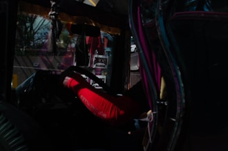 Inside a vehicle, a person wearing a red sleeve is holding the steering wheel. The interior is dimly lit with reflections and decals visible on the glass. Some text signs are present, along with a road and other vehicles in the background through a window.