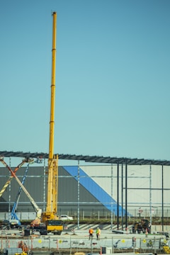 A construction site with a large yellow crane towering over two workers in safety gear. Steel beams are being erected for a new structure, and various construction vehicles and materials are present. The building in progress features panels with blue and gray colors.