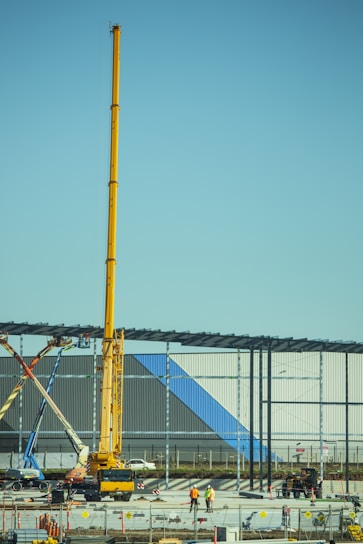 A construction site with a large yellow crane towering over two workers in safety gear. Steel beams are being erected for a new structure, and various construction vehicles and materials are present. The building in progress features panels with blue and gray colors.