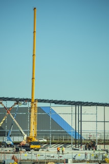A construction site with a large yellow crane towering over two workers in safety gear. Steel beams are being erected for a new structure, and various construction vehicles and materials are present. The building in progress features panels with blue and gray colors.