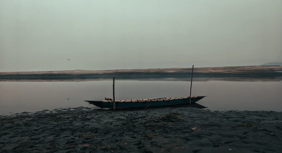 The image depicts a serene scene at a riverbank, with a small boat moored on the shore. The boat is painted in shades of blue and white, contrasting against the muted tones of the surrounding environment. It's positioned near the bank, facing towards the right side of the image. In the background, there are trees lining the riverbank, adding to the tranquil atmosphere. Above them all, a cloudy sky stretches out, casting a soft light over the scene.
