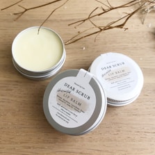 Several tins of Dear Scrub Unscented Lip Balm are placed on a wooden surface. One tin is open, revealing the lip balm inside. The labels on the tins provide information about ingredients and usage. Dried twigs are artistically arranged around the products, adding a natural element to the composition.