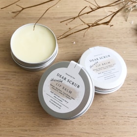 Several tins of Dear Scrub Unscented Lip Balm are placed on a wooden surface. One tin is open, revealing the lip balm inside. The labels on the tins provide information about ingredients and usage. Dried twigs are artistically arranged around the products, adding a natural element to the composition.