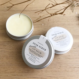 Several tins of Dear Scrub Unscented Lip Balm are placed on a wooden surface. One tin is open, revealing the lip balm inside. The labels on the tins provide information about ingredients and usage. Dried twigs are artistically arranged around the products, adding a natural element to the composition.