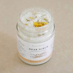 A glass jar labeled 'Dear Scrub Calendula Chamomile' filled with coarse sea salt and dried calendula flowers is placed on a light beige surface.