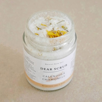 A glass jar labeled 'Dear Scrub Calendula Chamomile' filled with coarse sea salt and dried calendula flowers is placed on a light beige surface.