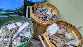 Several trash bins, mostly filled with various types of garbage like plastic bags, leaves, cardboard, and food containers. The bins are different colors, including green and yellow, and are located against a pale wall.
