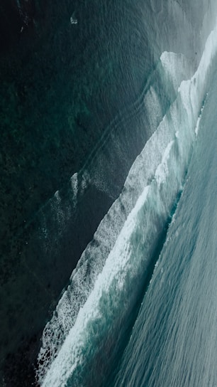 Waves crash against each other in the open ocean, creating a dynamic and textured pattern. The water varies in color from deep teal to lighter shades as the waves rise and fall.