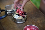 A close-up view of a person picking dried fruit from a small, open metal tin containing a mix of snacks including dried fruits and pretzels on a wooden table.