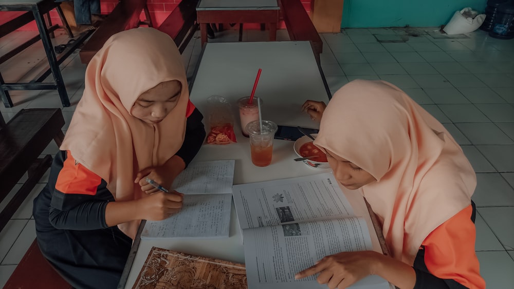 two girls sitting at a table with a book and a drink