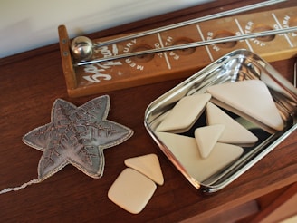 A wooden table holds a decorative object shaped like a snowflake next to a wooden board game with metal sliders. A metallic tray contains several smooth, cream-colored geometric shapes.