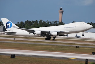 A large white airplane with the phrase 'Challenge accepted' on its side is taking off from a runway. The aircraft has a distinctive blue and white logo on the tail. In the background, there are green trees and a tall control tower.