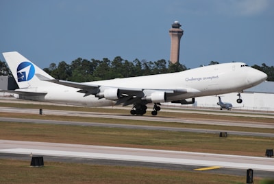A large white airplane with the phrase 'Challenge accepted' on its side is taking off from a runway. The aircraft has a distinctive blue and white logo on the tail. In the background, there are green trees and a tall control tower.