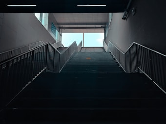 A spacious interior setting featuring a wide staircase leading upwards towards a brightly lit exit. The symmetrical railings on either side guide towards the outdoors, where daylight filters in. The subdued lighting indoors contrasts with the bright daylight outside, creating a strong sense of directionality and openness.