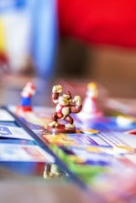 A close-up view of a board game featuring small figurines, including a monkey figure in the center wearing a red tie, with other characters visible in the background slightly out of focus. The board is colorful and detailed, with various sections and texts.