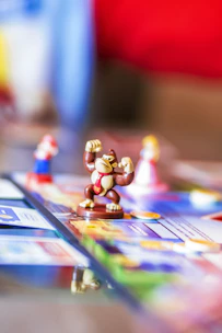 A close-up view of a board game featuring small figurines, including a monkey figure in the center wearing a red tie, with other characters visible in the background slightly out of focus. The board is colorful and detailed, with various sections and texts.