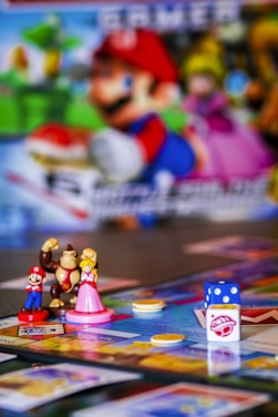 A colorful board game setup featuring small figurines of video game characters, a board with various spaces, plastic coins, and a dice. The background includes a blurred image of characters from the same video game franchise.