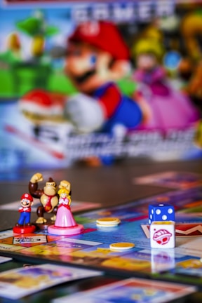 A colorful board game setup featuring small figurines of video game characters, a board with various spaces, plastic coins, and a dice. The background includes a blurred image of characters from the same video game franchise.