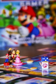 A colorful board game setup featuring small figurines of video game characters, a board with various spaces, plastic coins, and a dice. The background includes a blurred image of characters from the same video game franchise.