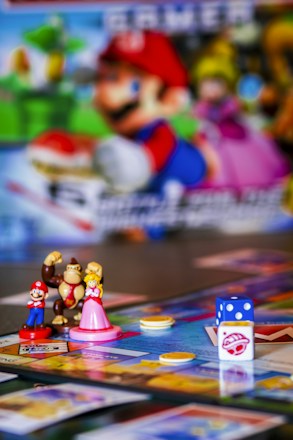 A colorful board game setup featuring small figurines of video game characters, a board with various spaces, plastic coins, and a dice. The background includes a blurred image of characters from the same video game franchise.