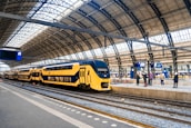 A modern yellow and blue double-decker train is stationed at a large, airy train station with a high, curved glass and metal roof. Several people are standing on the platform, some looking at the digital departure boards displaying information.
