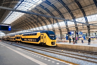 A modern yellow and blue double-decker train is stationed at a large, airy train station with a high, curved glass and metal roof. Several people are standing on the platform, some looking at the digital departure boards displaying information.