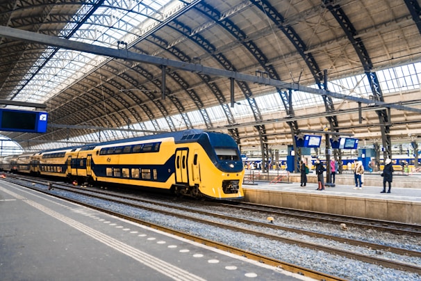 A modern yellow and blue double-decker train is stationed at a large, airy train station with a high, curved glass and metal roof. Several people are standing on the platform, some looking at the digital departure boards displaying information.