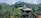 A house is situated amidst lush greenery with dense trees and palm trees around. The structure has a sloped roof with red shingles and appears to be partially constructed or undergoing renovation. In the background, a hill rises under a partly cloudy sky.